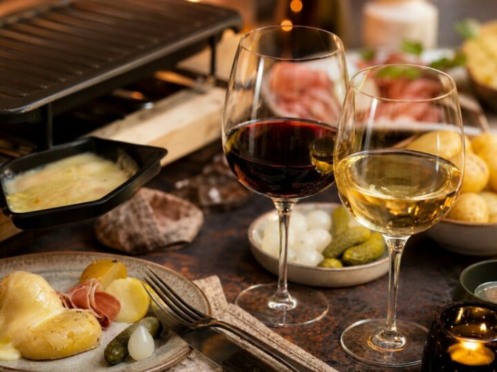 Zwei Gläser Rot- und Weißwein vor Raclette-Tisch mit Beilagen.