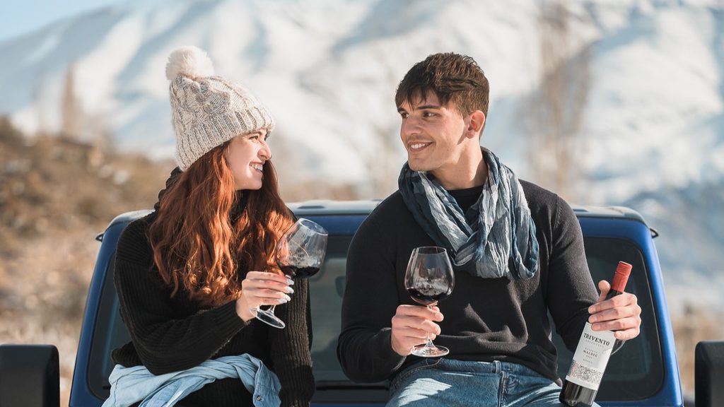 Ein junges Paar sitzt im Winter mit Wein auf der Motorhaube eines Geländefahrzeugs.